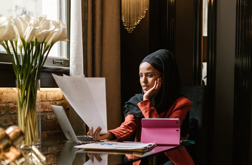 A woman in business attire with a hijab reviews documents while sitting at a laptop in a luxurious office setting.